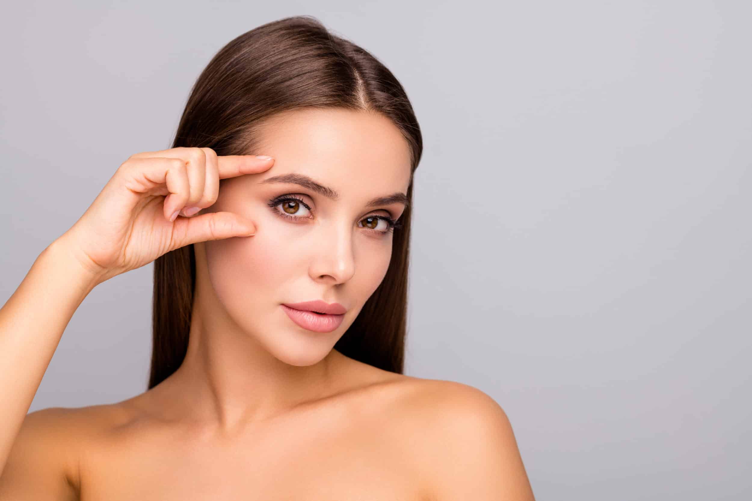 Close-up photo of a beautiful woman with youthful appearance, gently touching her eyebrow to highlight the effects of an endoscopic brow lift, isolated on a grey background