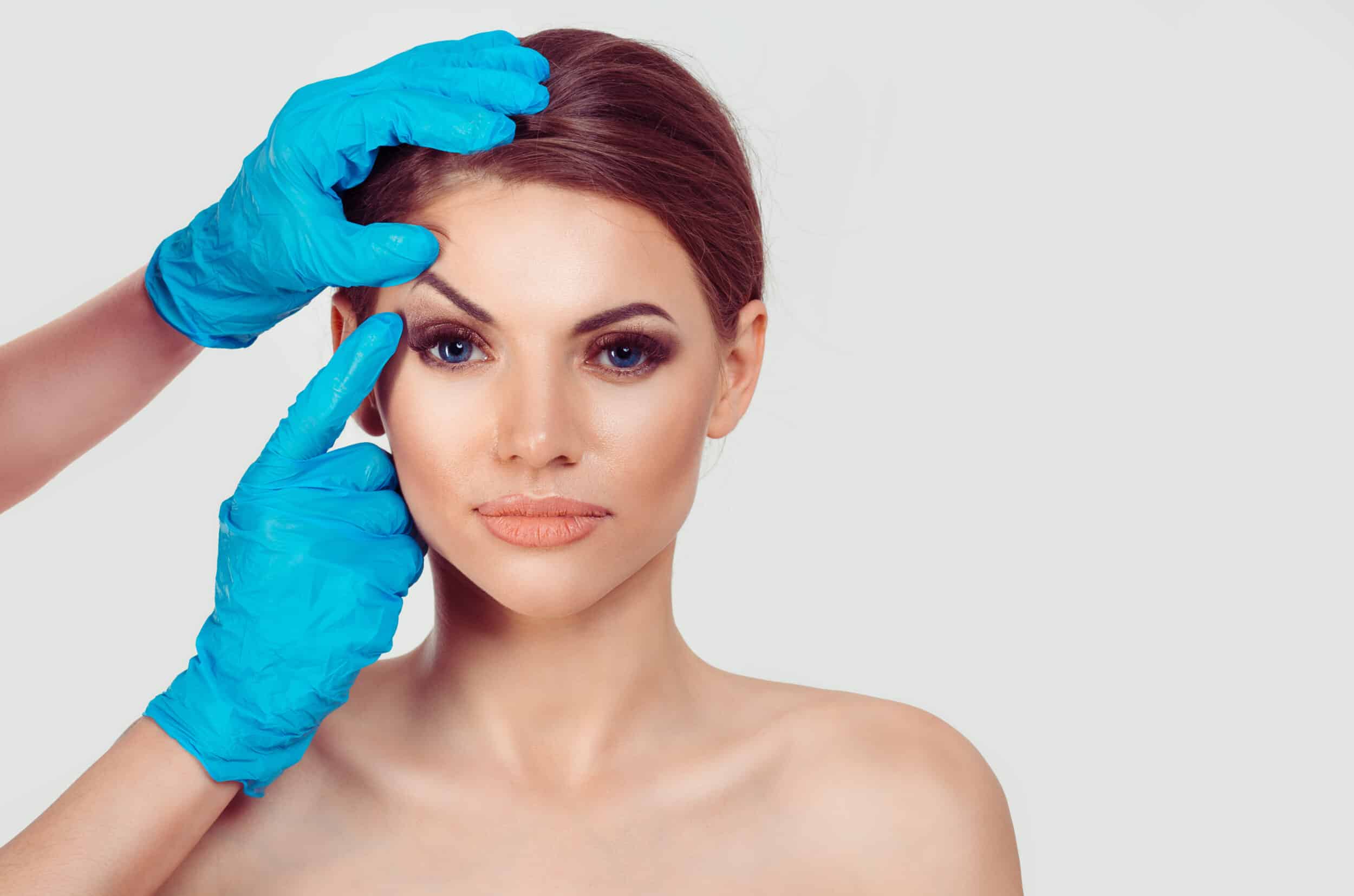Beautiful middle-aged woman preparing for upper eyelid blepharoplasty and endoscopic brow lifting, as a doctor in blue gloves points to her eye, highlighting cosmetic surgery for a youthful appearance against a white background